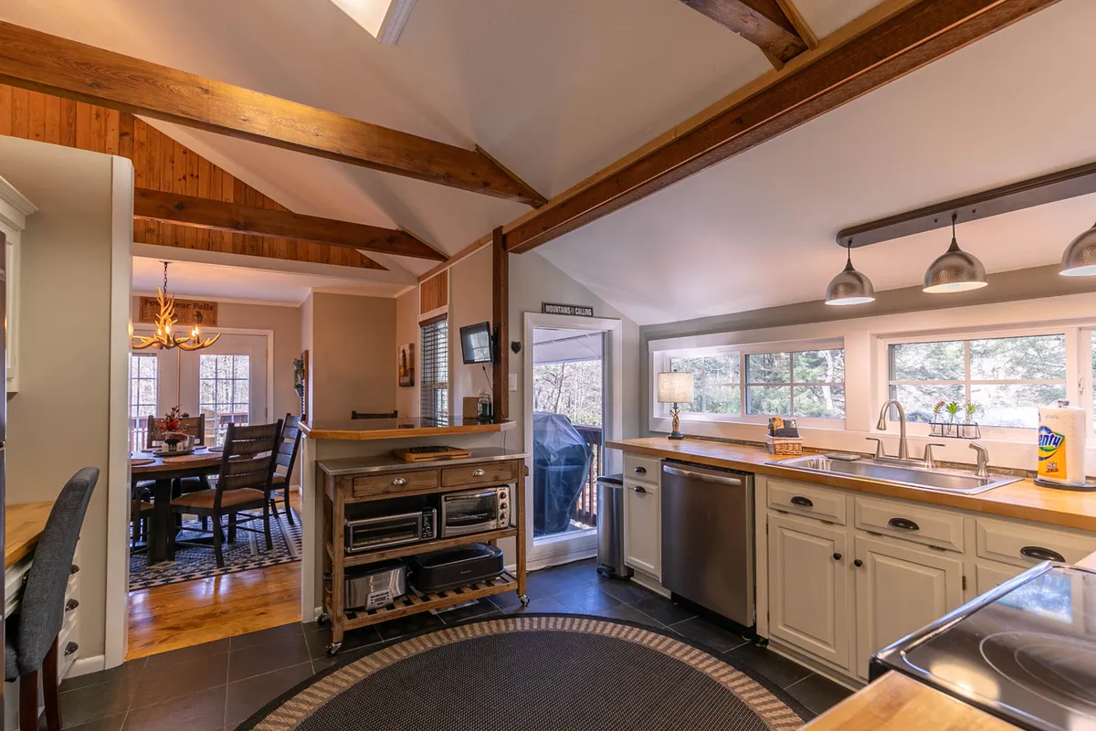 A view encompassing both the kitchen island and dining area, framed by large windows and wooden beams. The open-concept design shows the fluid connection between food preparation and communal dining, with natural light enhancing the inviting atmosphere.