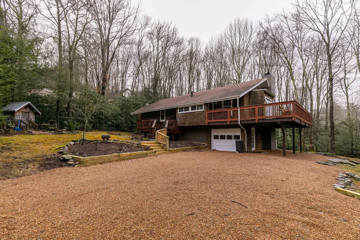 This exterior image showcases the house, a spacious deck, and the gravel driveway set against a backdrop of trees. The view suggests generous outdoor space perfect for families to gather, play, and enjoy nature together. The deck area hints at a welcoming environment for outdoor meals or relaxation.
