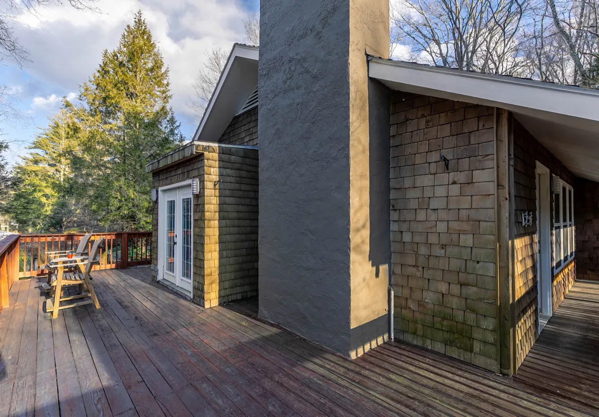 A spacious wooden deck attached to the house, set against a backdrop of trees. Large windows and a chimney add architectural interest, while the space is arranged for socializing, relaxation, and outdoor dining—ideal for barbecues or evening gatherings.