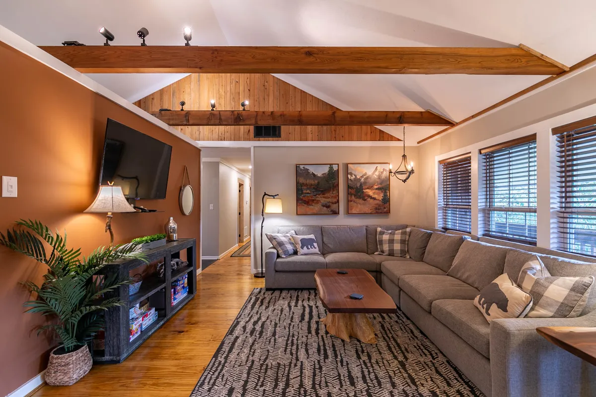 A large, plush sectional sofa faces a wooden coffee table and a wall-mounted TV, surrounded by decorative artwork and plants. The open and airy layout, with ample seating, is perfect for families or groups wanting to unwind or enjoy a movie night together. The overall ambiance is bright and welcoming, enhanced by natural light.