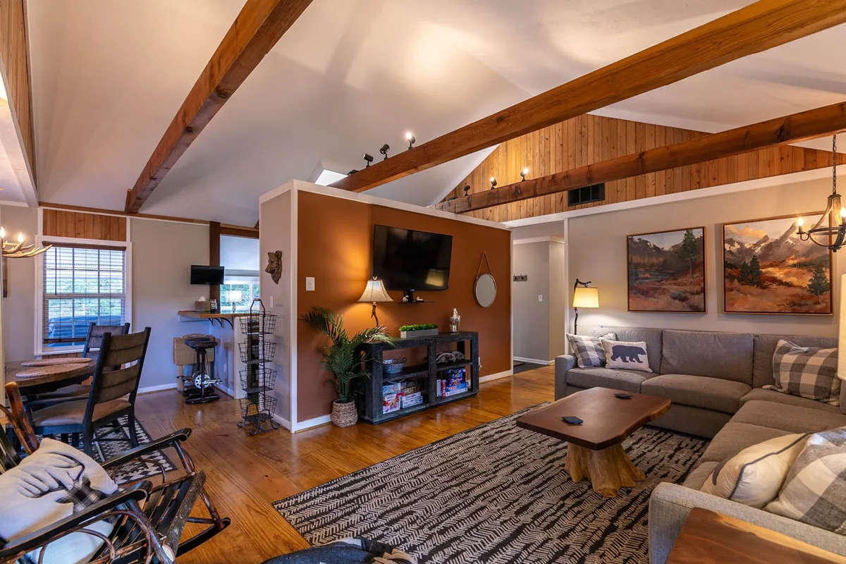 A cozy living space featuring exposed wooden beams, a soft sofa, a coffee table, decorative wall paintings, and a television. The room is filled with natural light, creating a cheerful, welcoming setting ideal for both everyday use and special occasions.