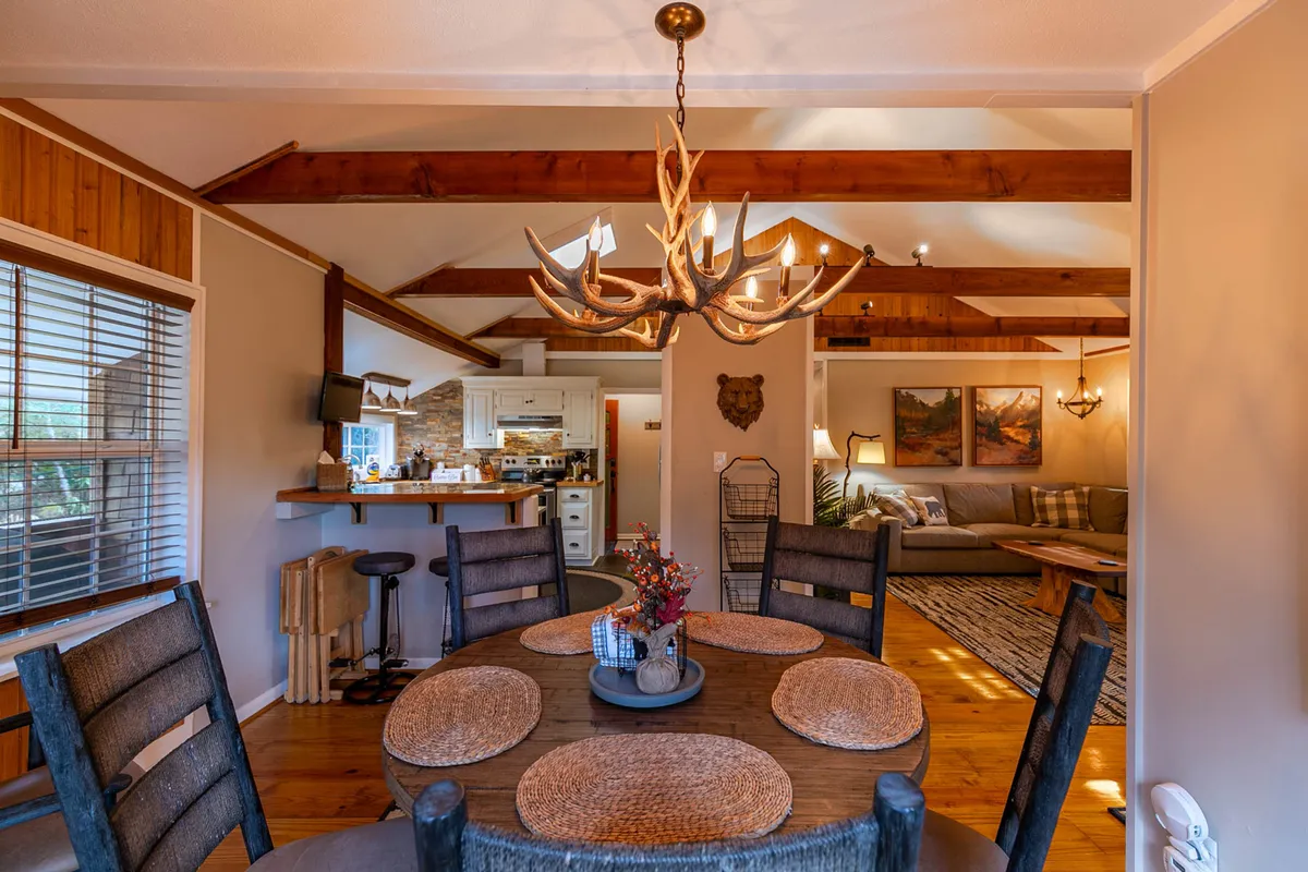 A warmly lit dining area with a rustic chandelier above a wooden table, adjacent to a cozy living room with a comfortable couch. The kitchen area is visible, creating a sense of openness and togetherness.