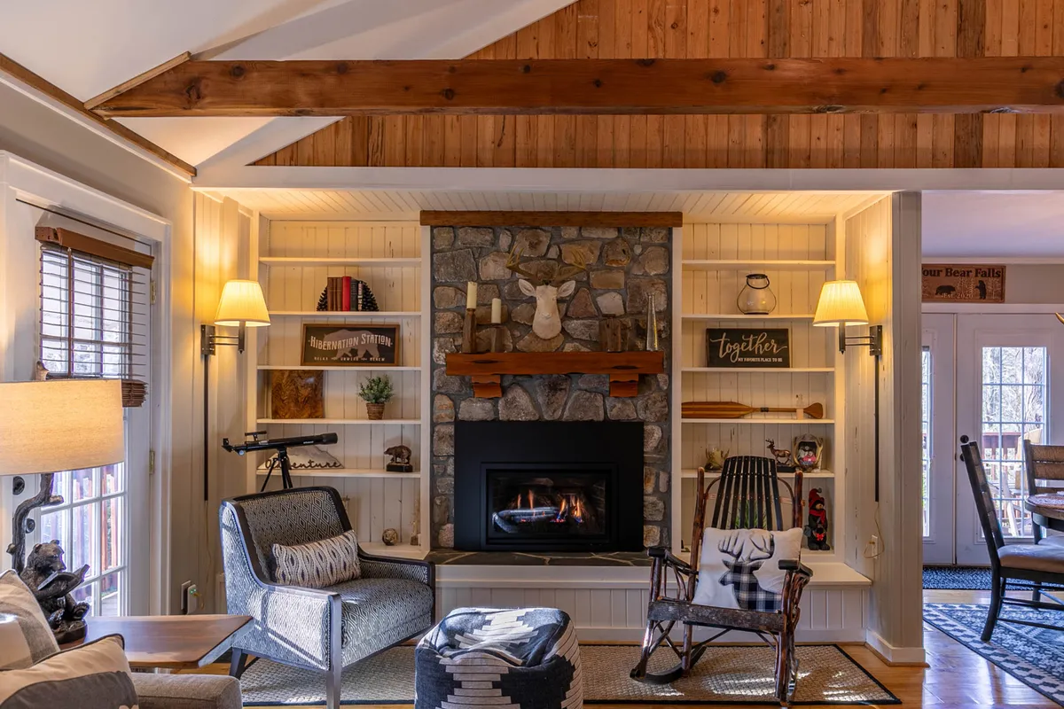 This image shows a spacious living room with a striking stone fireplace as the centerpiece, complemented by exposed wooden beams overhead. Comfortable seating invites guests to gather and relax, while shelves with tasteful mountain-inspired decor add character. Soft, warm lighting creates an inviting atmosphere, perfect for group conversations or quiet relaxation.