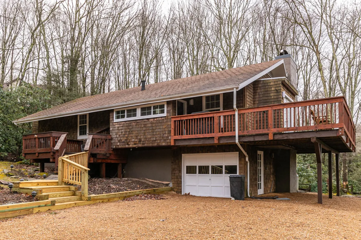 The image showcases the exterior of a charming single-family house surrounded by lush trees, featuring a spacious wooden deck perfect for gatherings. The inviting staircase leads up to the deck, and a garage door is visible, offering convenient parking. The setting is tranquil, with the natural beauty of Banner Elk and the Blue Ridge Mountains in the background, suggesting opportunities to relax with sunset views or enjoy a morning coffee outdoors.