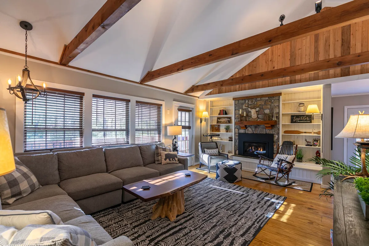 A warm, inviting living area featuring a stone fireplace, plush sofa, exposed wooden beams, and built-in bookshelves. The space is ideal for relaxing after a day outdoors, with seating arranged for cozy gatherings and a large rug adding comfort.
