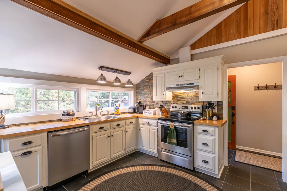 A spacious kitchen with a central island, stainless steel appliances, large windows letting in natural light, and exposed wooden beams overhead. The setting exudes warmth and invites both cooking and gathering, with plenty of counter space visible and a clear connection between kitchen and adjacent spaces.