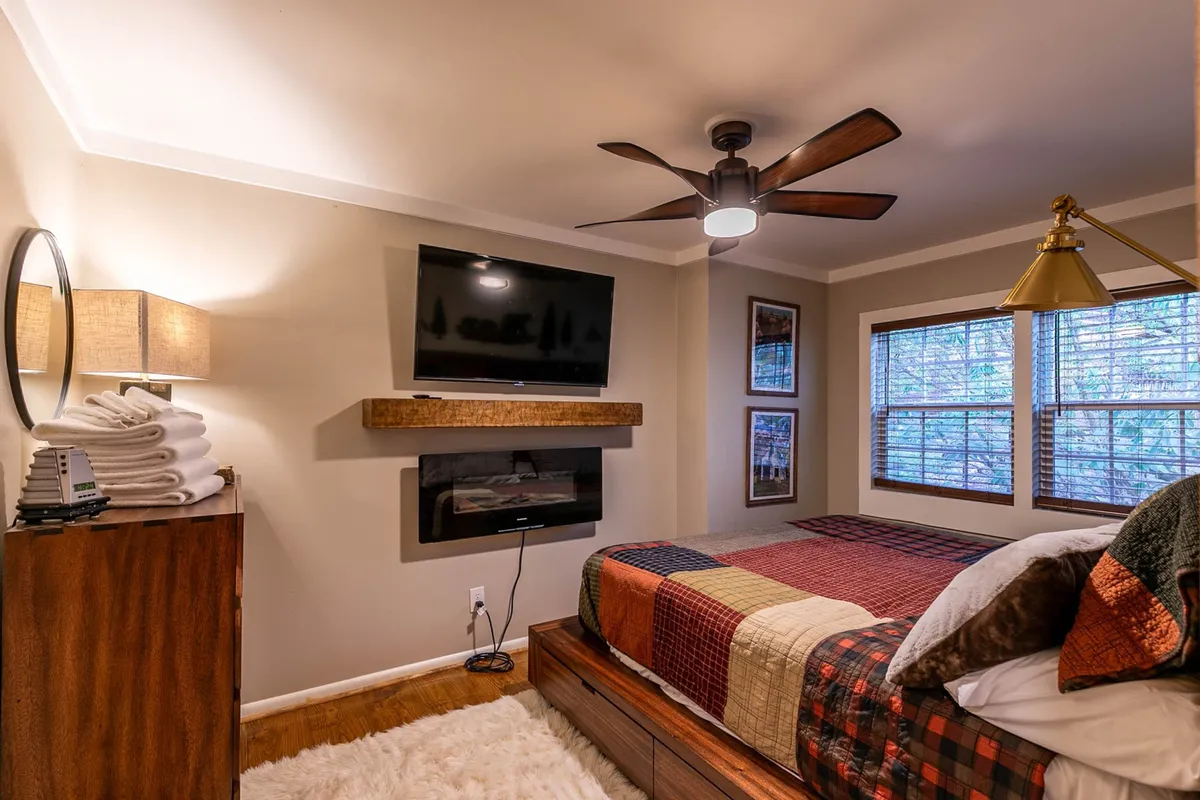 This image showcases a cozy bedroom featuring a bed adorned with a patterned quilt, a wall-mounted flat screen TV, a decorative lamp, a warm fireplace, and windows offering relaxing views. The space exudes warmth and comfort, with visually appealing details that invite rest and relaxation.