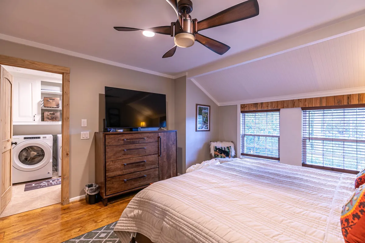 A bright, inviting bedroom with a bed dressed in a crisp white quilt, a large TV atop a dark wooden dresser, and a window letting in morning light through blinds. The space feels homely and comfortable, ideal for waking refreshed or enjoying quiet moments.